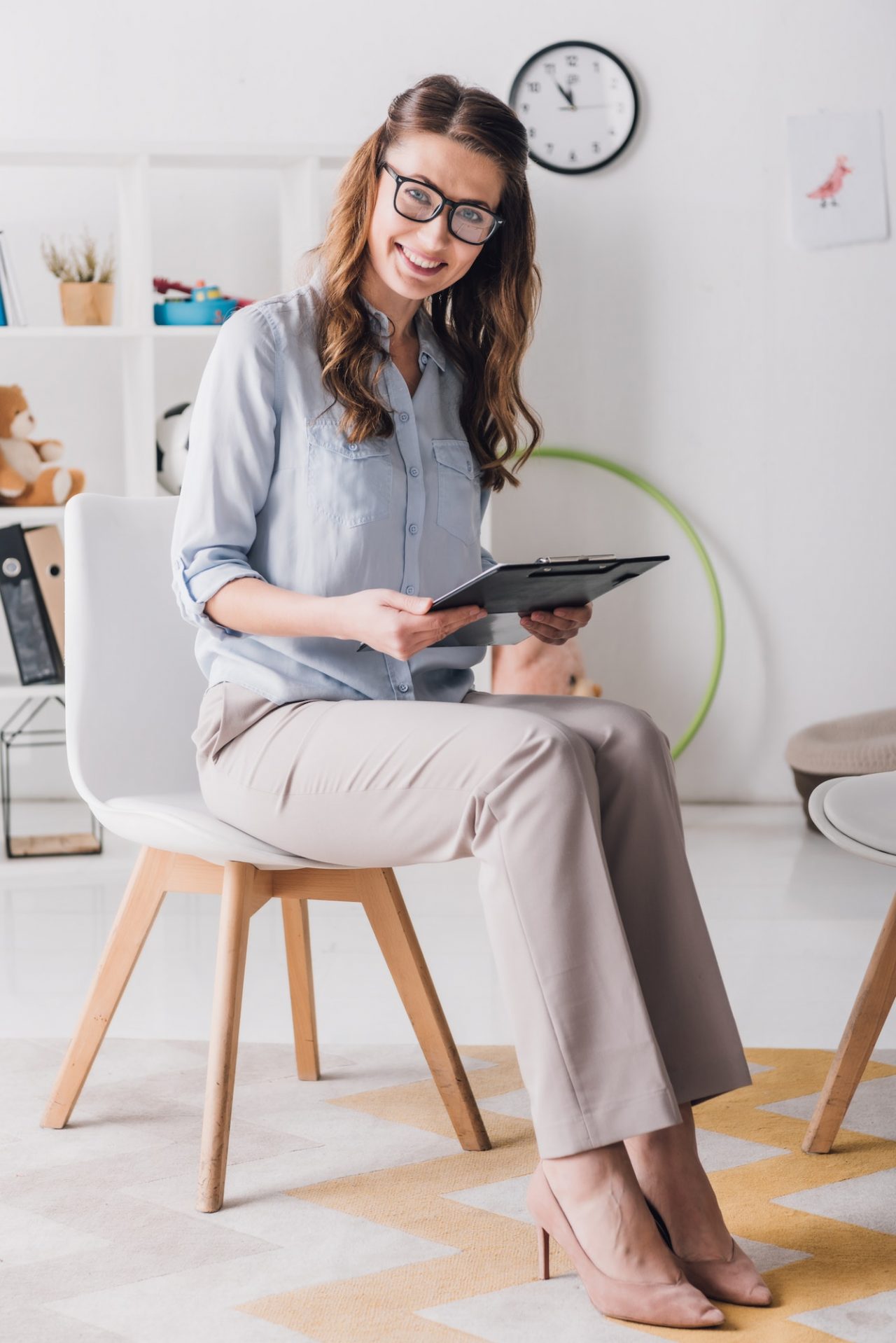 Smiling child psychologist with clipboard sitting on chair in office and looking at camera.jpg